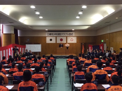 「三郷市少年消防クラブ入団式」と書かれた横断幕の下の檀上に立つ市長の話を聞いているオレンジ色のお揃いの上着を着て着席している少年消防クラブ員達の後ろ姿の写真