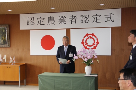 「認定農業者認定式」と書かれた横幕と国旗と三郷市旗が飾られた室内で挨拶を行っている市長の写真