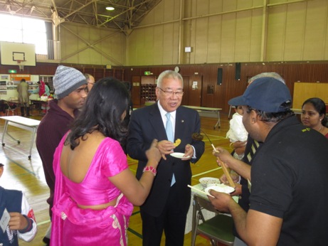 外国人に囲まれた市長が小さな紙皿に入ったお菓子を食べている様子の写真
