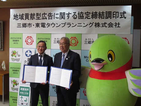 「地域貢献型広告に関する協定締結調印式」と書かれたポスターが貼られている三郷市のボードの前で、三郷市長と東電タウンプランニング株式会社の関係者が協定書を手に持ち、かいちゃんとともに記念撮影をしている写真