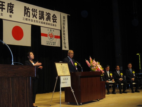 「防災講演会」と書かれた横断幕と国旗・三郷市自主防災組織連絡協議会の旗が吊るされているステージ上で、赤・ピンク・黄色・白の豪華な花々で彩られた花台と並ぶ演台に立ち挨拶の言葉を述べる三郷市長とスタンドに置かれた表彰状のそばに立ち手話通訳をする女性、ステージ上に並べられてたパイプ椅子に座り話を聞く胸章を付けた来賓者の写真