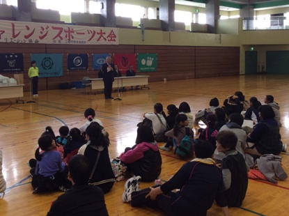 「第25回チャレンジスポーツ大会」と書かれた横断幕の下に設置されたマイクスタンドの前に立っている市長の話を床に座って聞いている参加者の後ろ姿の写真