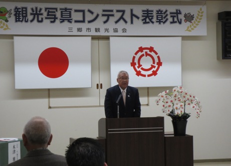 「観光写真コンテスト表彰式」と書かれた看板と、日の丸、三郷市の旗が掲げられた壁を背に、演台に立って挨拶をする市長の写真