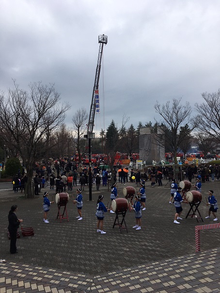「祝 謹賀新年」と書かれた垂れ幕が高く上げられた梯子に掛けられ、手前の広場に設置された5つの大太鼓を2名1組の法被を着た中学生が演奏を行っている様子の写真