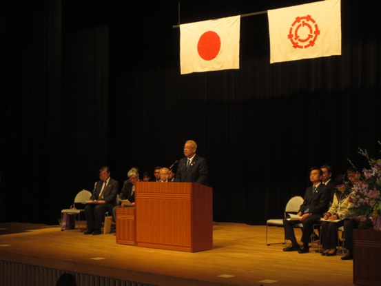 日の丸と三郷市の旗が掲げられた舞台の上で、着席している関係者たちを背に、演台に立って話をする市長の写真