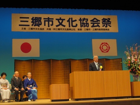「三郷市文化協会祭」と書かれた横断幕と国旗と三郷市旗に金屏風が設置されたステージの演壇で挨拶を行っている市長の写真