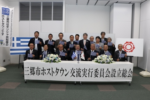 「三郷市ホストタウン交流実行委員会設立総会」と書かれた横幕が2つの長机を繋げた前に貼られ、前列に座っている市長を含め6名と、後列の10名の方々が小さなギリシャの国旗を持ち並んでいる記念写真