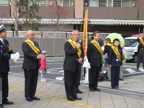 駐車スペース横の歩道で、黄色いタスキをかけ、マイクを持って話をしている市長と、その両側に同じタスキをかけて並ぶ関係者、後方のオレンジ色ののぼりの横に立つかいちゃんを写した写真