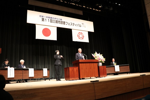 「第11回三郷市読書フェスティバル」と書かれたボードと国旗・三郷市旗が吊るしてあるステージ上で、ピンク・黄色・白の豪華な花々で彩られた花台と並ぶ演台に立ち挨拶の言葉を述べる三郷市長と演台の横で手話通訳をしている女性、ステージ上に配置された席に座り話を聞く胸章を付けた来賓者の写真
