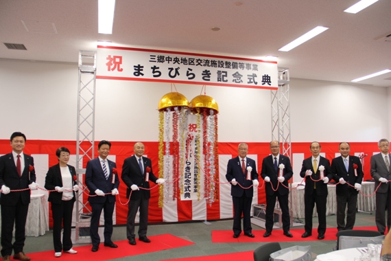 「祝 三郷中央地区交流施設整備等事業まちびらき記念式典」と書かれた横断幕と壁に紅白幕が貼られた会場で、市長を含め左右に9名の関係者が並び横断幕の下のくす玉が開いた様子の写真