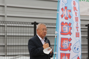 水道週間と書かれたのぼり旗の手前でマイクを右手に持ち公園の蛇口点検プロジェクト出発式で挨拶を行っている市長をアップで撮影した写真
