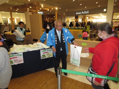 水色の法被を着た市長が小松菜が入った白色の袋を右側の赤色の服を着た女性に手渡している様子の写真