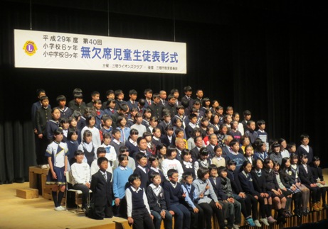 「平成29年度第40回小学校6ヶ年、小・中学校9ヶ年無欠席児童生徒表彰式」と書かれた横断幕が吊り下げられているステージ上で、受賞した134名の子どもたちが並んで三郷市長とともに集合写真を撮っている様子の写真