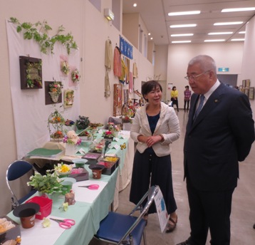 壁やクロスがかけられた長テーブルの上に様々なフラワーアートの作品が飾られている傍で女性の話を聞いている市長の写真