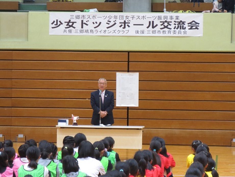 「少女ドッジボール交流会」の横断幕が2階の観客席から掛かった壁を背に、長机の後ろに立った市長が、整列して座っている子供たちに向かって話をしている写真