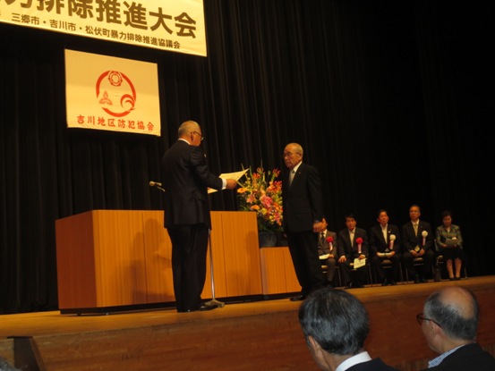 豪華な花々が飾られている花台と演台の前で、表彰状の内容を読み上げる三郷市長と向かい合って立つ功労者の男性の写真