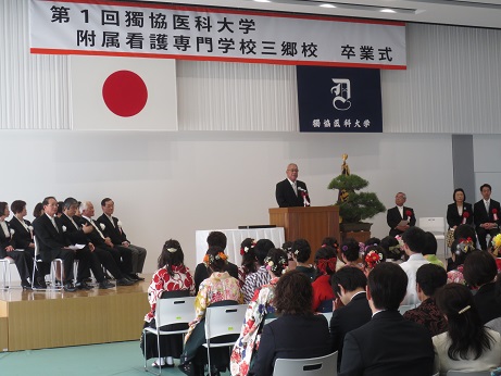 「第1回獨協医科大学附属看護専門学校三郷校卒業式」と書かれた横断幕と国旗・獨協医科大学の校章機が吊り下げられているステージ上で、立派な盆栽が置かれた花台と並ぶ演台に立ち挨拶の言葉を述べる三郷市長と、ステージ上に置かれた椅子に座る胸章を付けた来賓者、ステージを向いて並べられた椅子に座り話を聞く袴姿の卒業生の写真