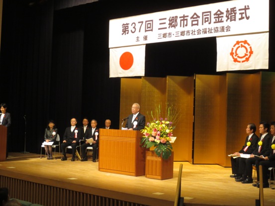 「第37回三郷市合同金婚式」と書かれた横断幕と国旗・三郷市旗が吊り下げられているステージ上で、赤・ピンク・黄色・白の豪華な花々で彩られた花台と並ぶ演台に立ち挨拶の言葉を述べる三郷市長と、ステージ上に置かれたパイプ椅子に座り話を聞く胸章を付けた来賓者の写真