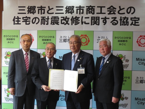 「三郷市と三郷市商工会との住宅の耐震改修に関する協定」と書かれたポスターが貼られている三郷市のボードの前で、三郷市長が三郷市商工会の会長と協定書を片方ずつ持ち合い、副会長と関係者の男性とともに記念撮影をしている写真