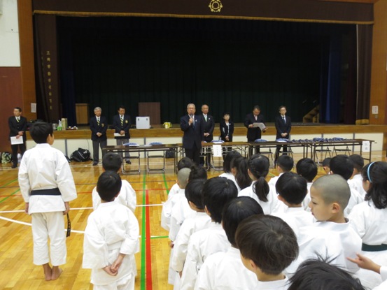 第25回近隣市町・市内交流空手道大会の開会式にて、前に立って話をする三郷市長と向かい合って列ごとに並んで立ち話を聞いている空手着を着た子供たちの写真