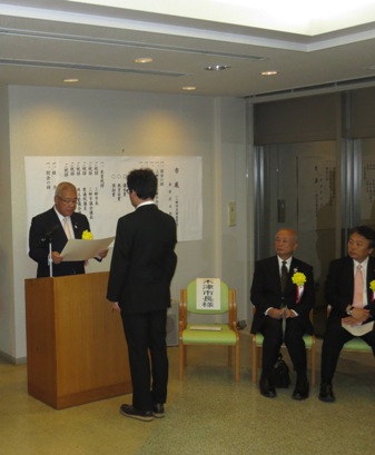 演台に立ち、正面に立つ受賞者を表彰している市長と、演台の向かって右側に置かれた椅子に座った来賓の写真