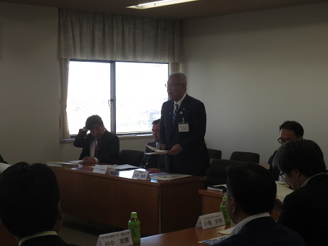 席を立ち資料を手に持ちながら挨拶の言葉を述べる三郷市長と席札とペットボトルのお茶が置かれている席に座り話を聞く参加者の写真