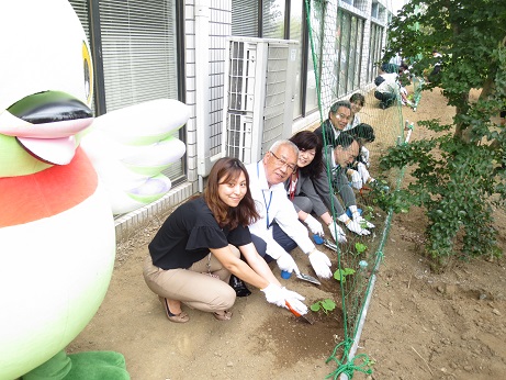 建物の真横にグリーンネットが張ってあり、ネットのすぐ下にスコップを使ってゴーヤの苗を植えている三郷市長と職員、立って見守るかいちゃんの写真