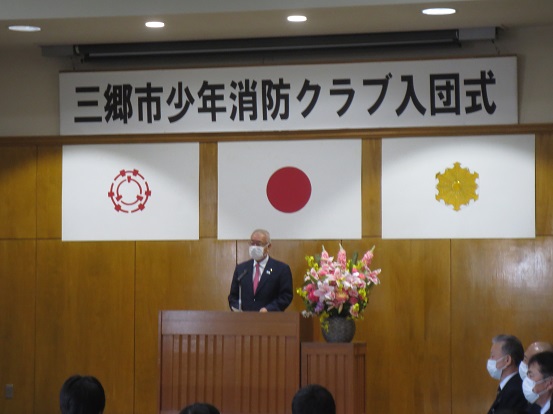 「三郷市少年消防クラブ入団式」と書かれた横断幕と三郷市章、国旗、消防紋章が掲げられた会場で挨拶をしている三郷市長と、席に座り聞いている関係者の方々の写真