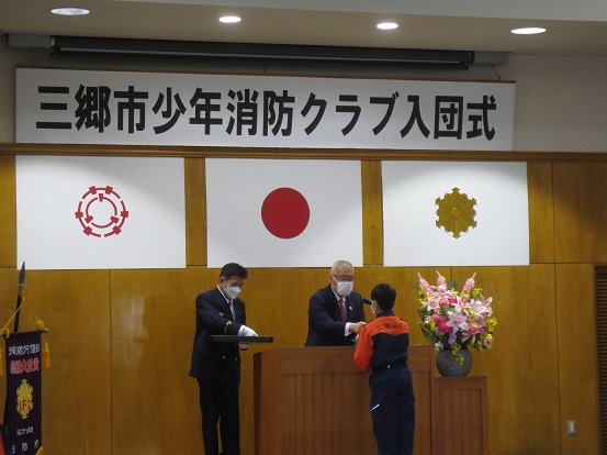 「三郷市少年消防クラブ入団式」と書かれた横断幕と三郷市章、国旗、消防紋章が掲げられた会場で、三郷市長がオレンジとネイビーの制服を着た方に少年消防手帳及びクラブ印章を交付している様子の写真