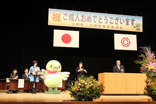 「祝ご成人おめでとうございます」と書かれたボードと国旗・三郷市旗が吊るしてあり、ステージ前方には色とりどりの豪華な式典花が飾られているステージ上で、演台に立ち挨拶の言葉を述べる三郷市長と、演台の横で手話通訳をしている女性、白いタスキをかけたかいちゃんと女性に抱きかかえられたつぶちゃんの写真