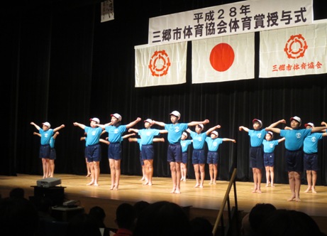 紅白帽と青い体操服を着用した子どもたちが両手を水平に伸ばし、ステージ上でラジオ体操の実演を行っている写真