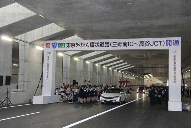 「東京外かく環状道路(三郷南インターチェンジ~市川高谷ジャンクション)開通」と書かれた特設の門を潜り抜ける白い車と、両脇から見守るたくさんの関係者たちの写真