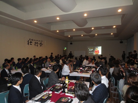会場のテーブルに料理が並んでいる部屋で、たくさんの関係者たちが正面のモニターに映る映像を見つめている写真