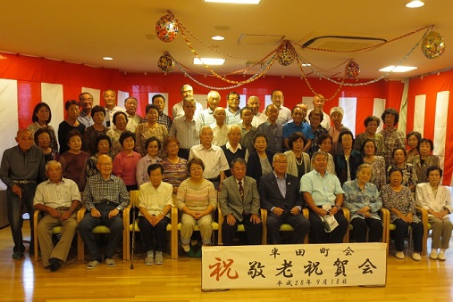 「半田町会 祝 敬老祝賀会」と書かれた横幕が立てかけられ、壁に沿って紅白幕が貼られた室内で、市長と参加者が並んでいる記念写真
