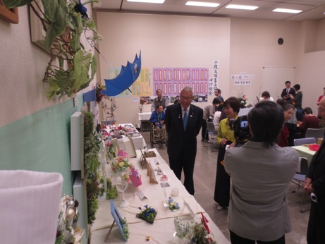 向かって左側の壁に蔦のウォールグリーンアートやリース、青い傘など、その下に置かれた白いカバーが掛けられたテーブルの上に、フラワーアレンジメントやフォトフレームなどの作品展示がされており、それらを鑑賞しながら談笑する三郷市長と女性、その様子をビデオカメラで撮影するカメラマンの写真