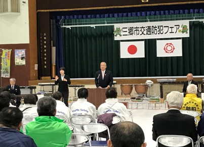 舞台に「三郷市交通防犯フェア」と書かれた横断幕と国旗、三郷市の旗が掲げられており、その舞台の下に設置された打楽器を背に、演台に立った市長が客席を向いて挨拶をしている様子と、隣で手話通訳をする女性、市長の方を見ている参加者たちを写した写真