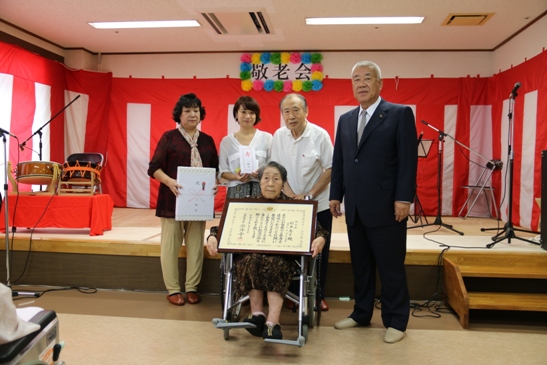 額縁に入った賞状を持って車いすに座っている鈴木島子さんと3名の家族と市長がステージの手前に並んでいる記念写真