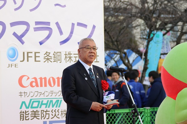 「みさとシティハーフマラソン」の文字と協賛企業の名前が書かれたボードの前に立ち、赤い胸章を付けた三郷市長が挨拶の言葉を述べている写真