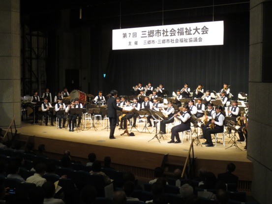 「第7回三郷市社会福祉大会」の吊り看板が設置されている舞台上で、吹奏楽部の生徒たちが演奏をしている写真
