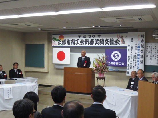 「平成30年三郷市商工会新春賀詞交歓会」と書かれた横断幕と国旗・三郷市商工会の紫色の旗がホワイトボードの前に吊るしてあり、赤・ピンク・黄色の豪華な花々で彩られた花台と並ぶ演台に立ち挨拶の言葉を述べる三郷市長と、白いカバーが掛けられている席に座り話を聞く胸章を付けた関係者の写真