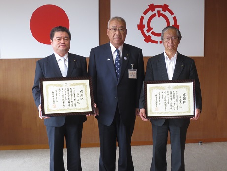 国旗と三郷市旗が吊り下げられている壁の前で、三郷市長と感謝状を持った男性2名がそれぞれ三郷市長の両隣に立っている記念写真