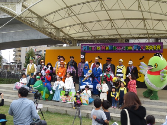 紫地にカラフルな文字で「MisatoHalloweenFes2018」と書かれたボードが貼り付けられているステージ上で、個性豊かなハロウィンのコスチュームを着用した表彰された参加者が並んで記念撮影をしている写真