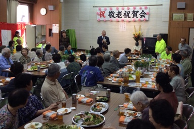 「敬老祝賀会」と書かれた大広戸東会館のステージの下で挨拶を行っている市長の話を飲み物や料理が並んだテーブルの席に座って聞いている参加者の写真