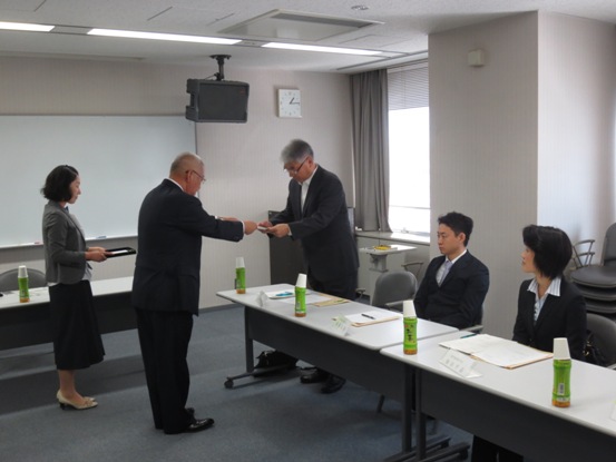 委託書を手渡す三郷市長と席を立って受け取る委員の男性、その様子を見守る介添人の女性と席に座る委員のメンバーの写真
