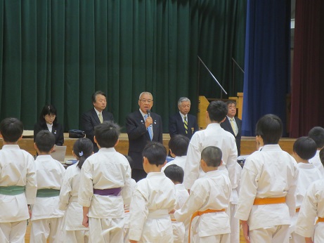 体育館のステージ前に立ち挨拶の言葉を述べる三郷市長と市長の横に並ぶ関係者、向かい合って立ち話を聞く空手着を着用し紫・緑・オレンジの帯を締めた子どもたちの写真