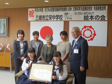 「祝平成29年度子どもの読書活動優秀実践校・団体文部科学大臣表彰受賞市長表敬訪問」と書かれたポスターと国旗・三郷市旗が吊るされた壁の前で、栄中学校の生徒2名が表彰状を一緒に持ってソファー椅子に座り、その後ろに三郷市長とライブラリーボランティア絵本の会のメンバーが立って記念撮影をしている写真