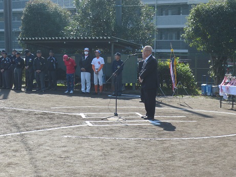 亀有信用金庫グラウンドに立ち挨拶の言葉を述べる三郷市長と、ベンチ近くに立ち話を聞くユニフォームを着用した関係者の写真