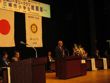横断幕と国旗、ロータリークラブの旗が掲げられた舞台で、演台に立って挨拶をする市長と、その後方の席に座るロータリークラブメンバーや来賓の写真