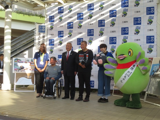 左から、銅メダルを首から掛けた三井梨紗子さん、花岡伸和さん、木津市長、越智貴雄さん、つぶちゃんのぬいぐるみを抱きかかえた三上大進さん、白いタスキをかけたかいちゃんが三郷市のバックスクリーンの前で記念撮影をしている写真
