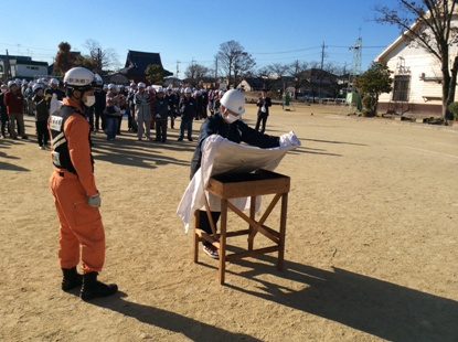 離れた場所で参加者が見ているなか、オレンジ色の作業着を着た消防隊員の指導のもと、ヘルメットを被った参加者が台の上に白色の布のような物を被せている初期消火訓練の様子の写真